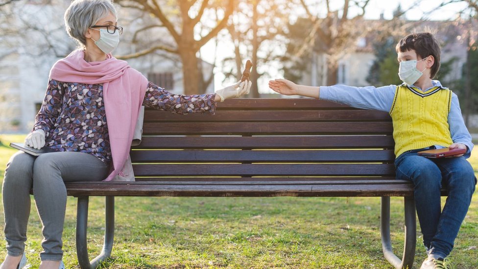 An older woman wearing a mask passes food to a boy on a bench, also wearing a mask