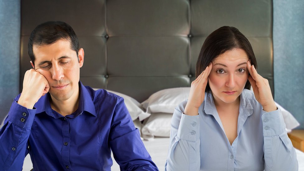 A worried woman and a bored man sitting on their bed.