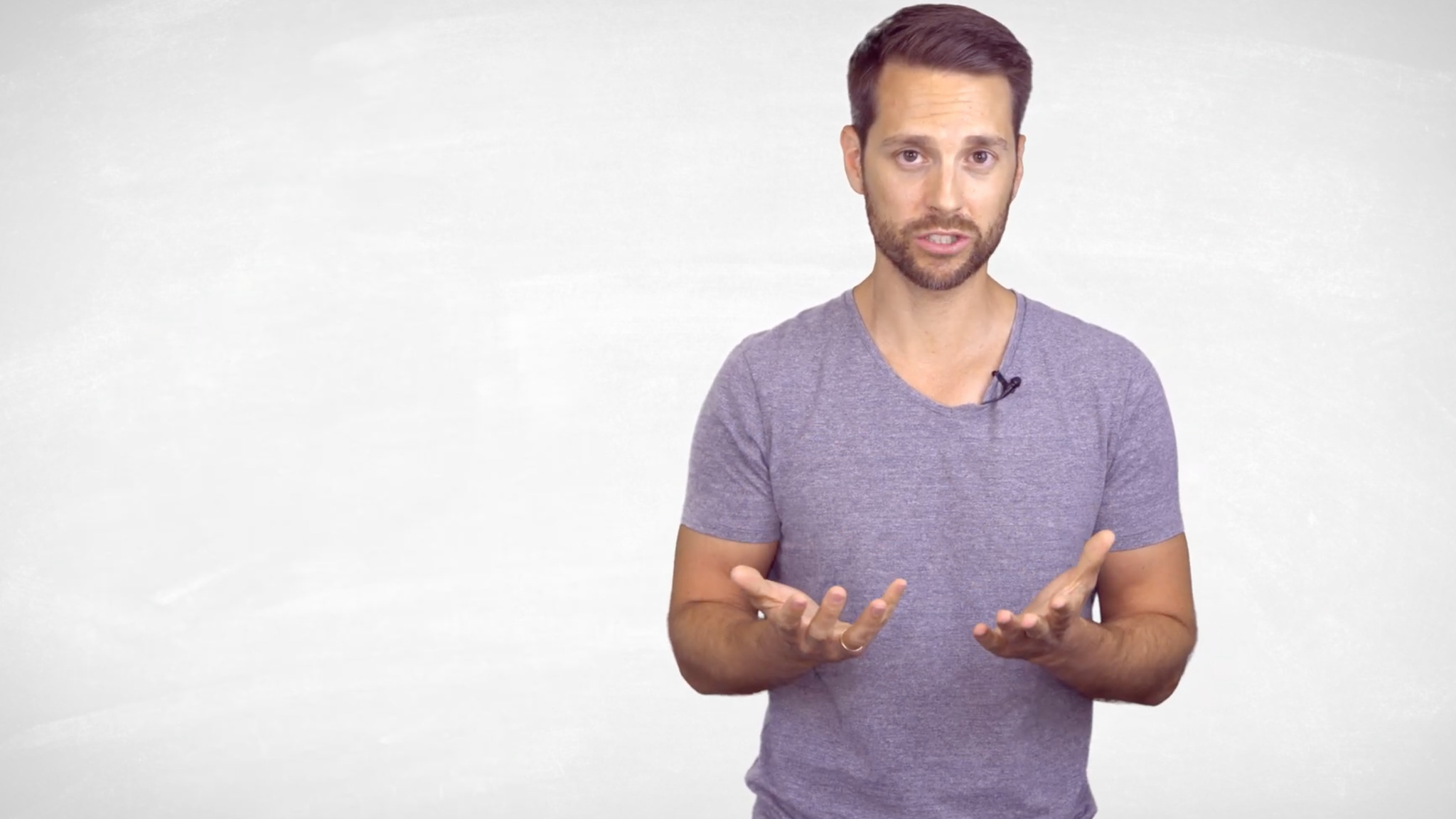 A man in a grey T-shirt stands in front of a grey screen addressing the camera as if talking